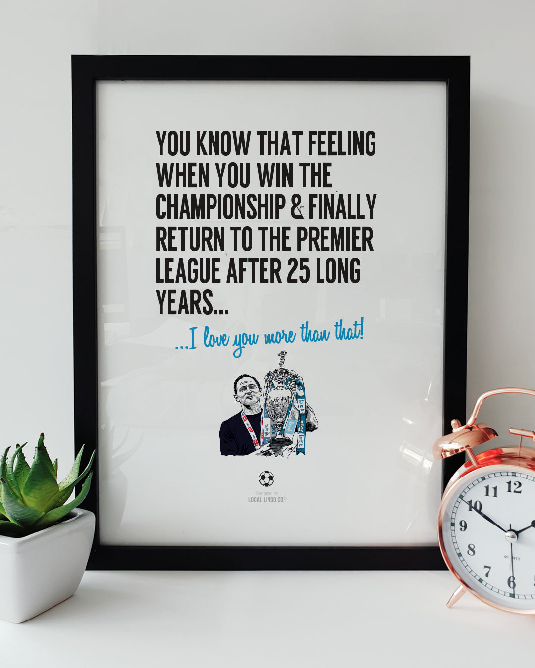 Framed football wall art celebrating Coventry City's promotion. Text reads: "You know that feeling when you win the Championship & finally return to the Premier League after 25 long years... I love you more than that!". Features a hand-drawn illustration of Frank Lampard with the Championship trophy. Displayed in a black frame next to a succulent and copper alarm clock. Designed by Local Lingo.