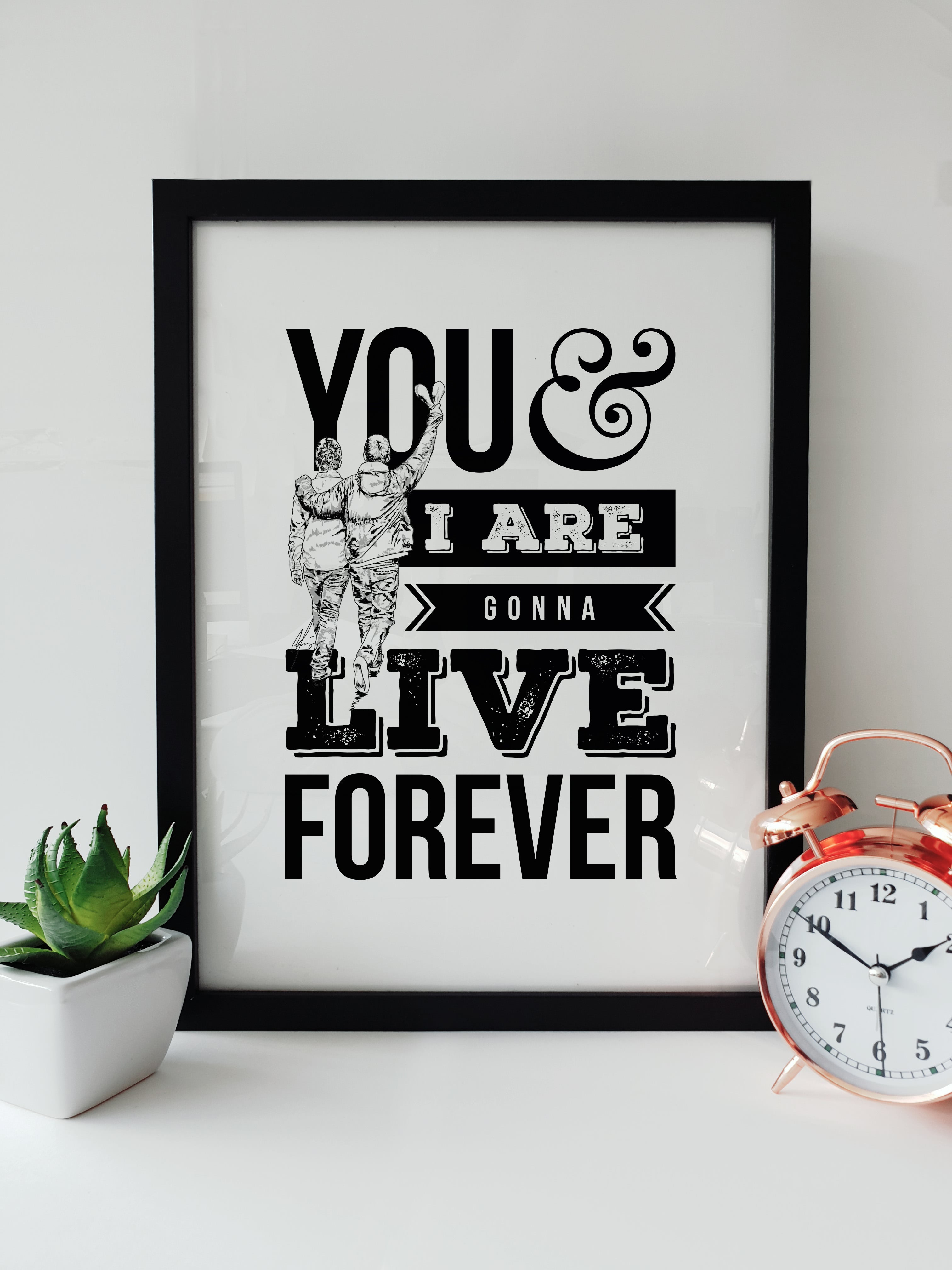 Framed black and white typographic print on a white shelf. The text reads "YOU & I ARE GONNA LIVE FOREVER" with an illustration of Liam and Noel Gallagher walking with their arms around each other. Designed by Local Lingo.