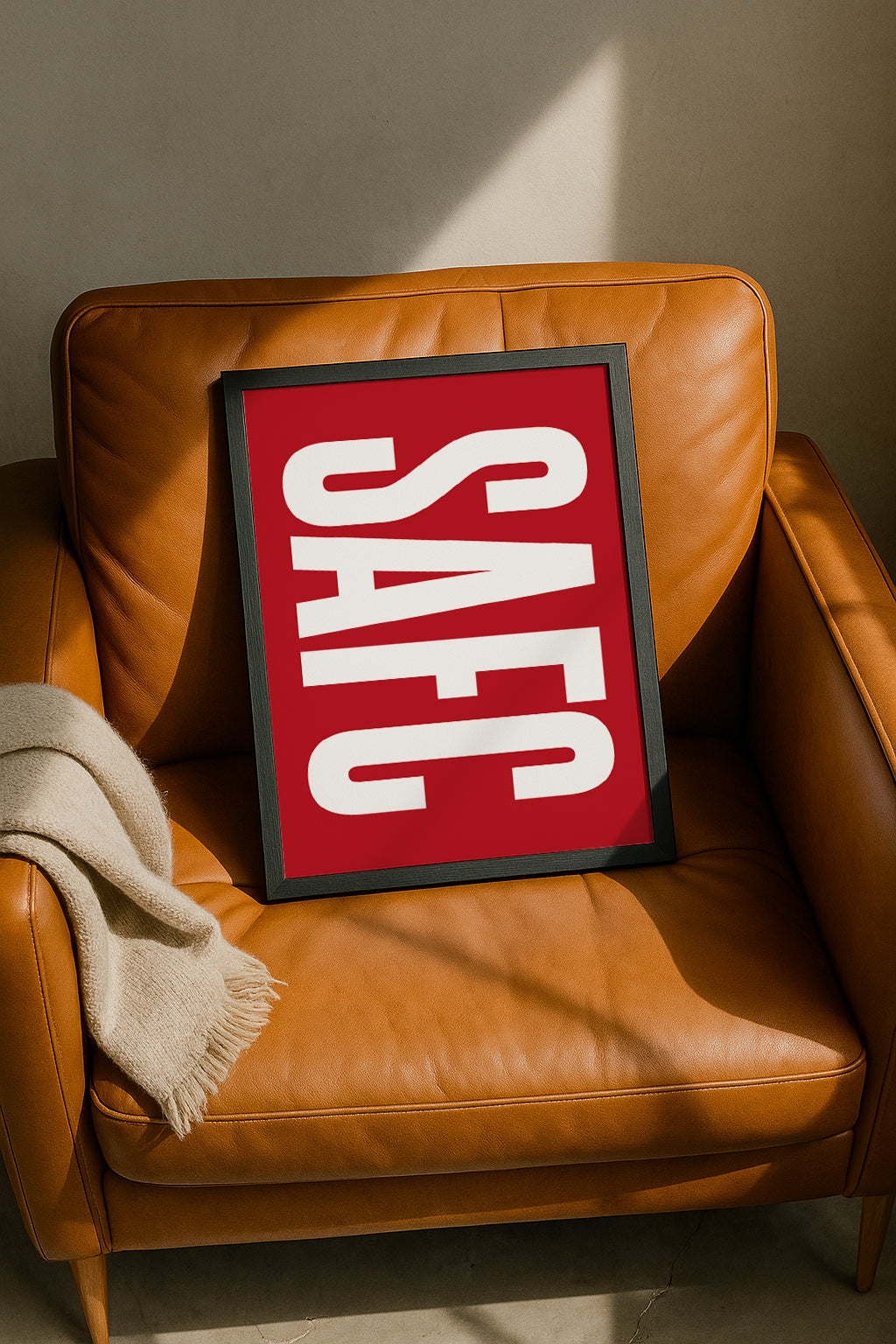 A framed SAFC typography art print resting casually on a tan leather armchair next to a beige throw blanket. The print features bold, retro-style white 'SAFC' lettering stretching vertically across a solid red background. Modern Sunderland football decor designed by Local Lingo.