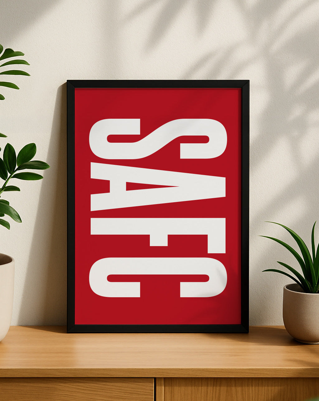 A framed SAFC typography art print displayed vertically on a light wooden sideboard, flanked by two small green potted plants with natural sunlight casting shadows on the wall behind it. The print features bold white 'SAFC' lettering on a solid red background. Minimalist Sunderland football wall art designed by Local Lingo.