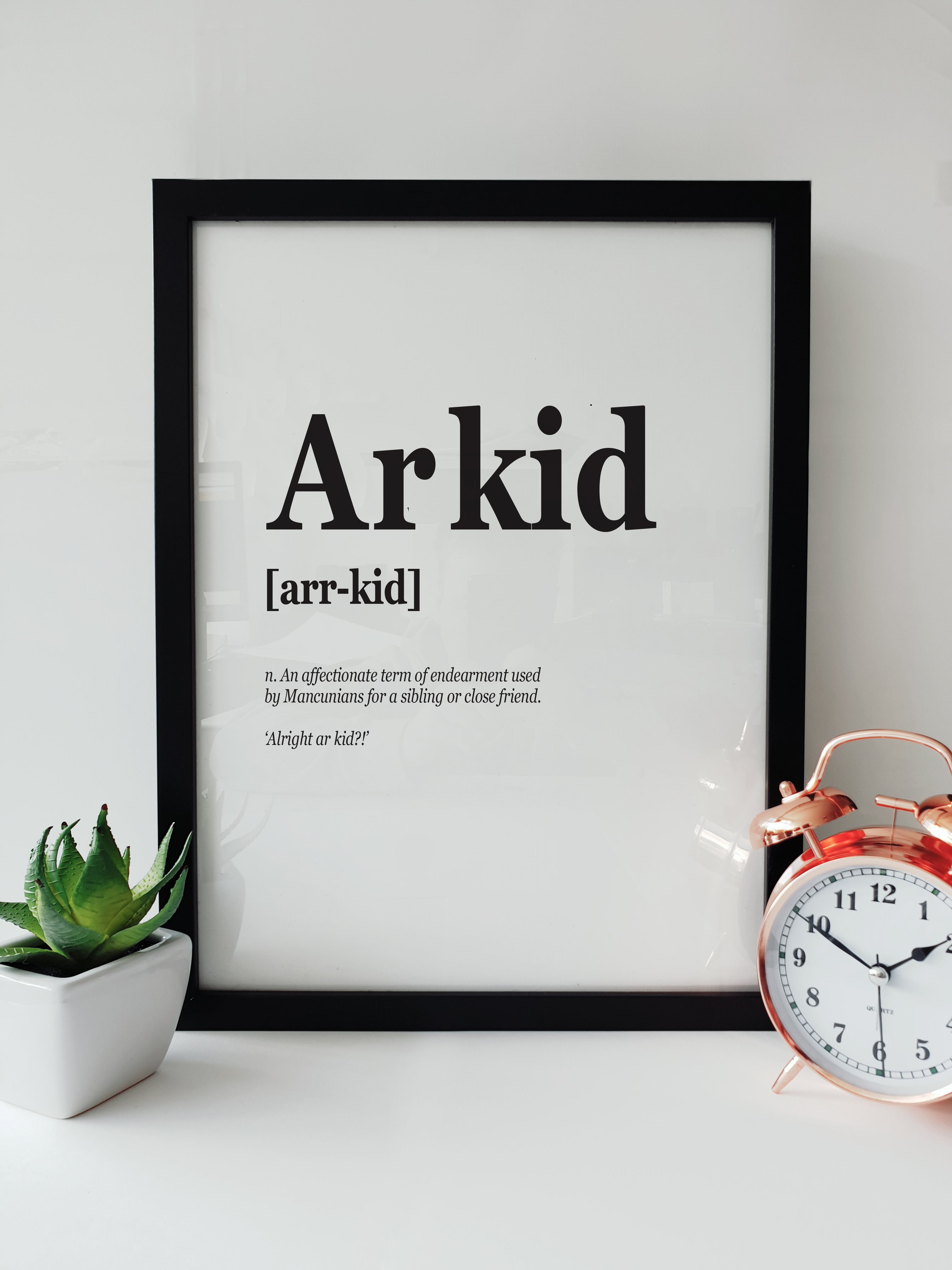 Framed typographic print on a white shelf titled "Arkid". The text defines the term as "An affectionate term of endearment used by Mancunians for a sibling or close friend." Displayed next to a small plant and a copper alarm clock. Designed by Local Lingo.