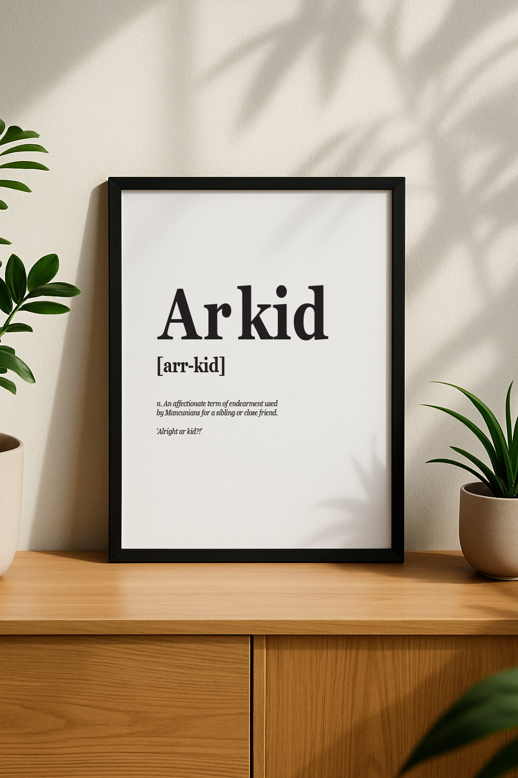 Vertical framed poster of the "Arkid" definition print hanging on a beige wall. The bold black text stands out against the clean white background, styled above a wooden sideboard with green plants for a modern home interior look. Designed by Local Lingo.