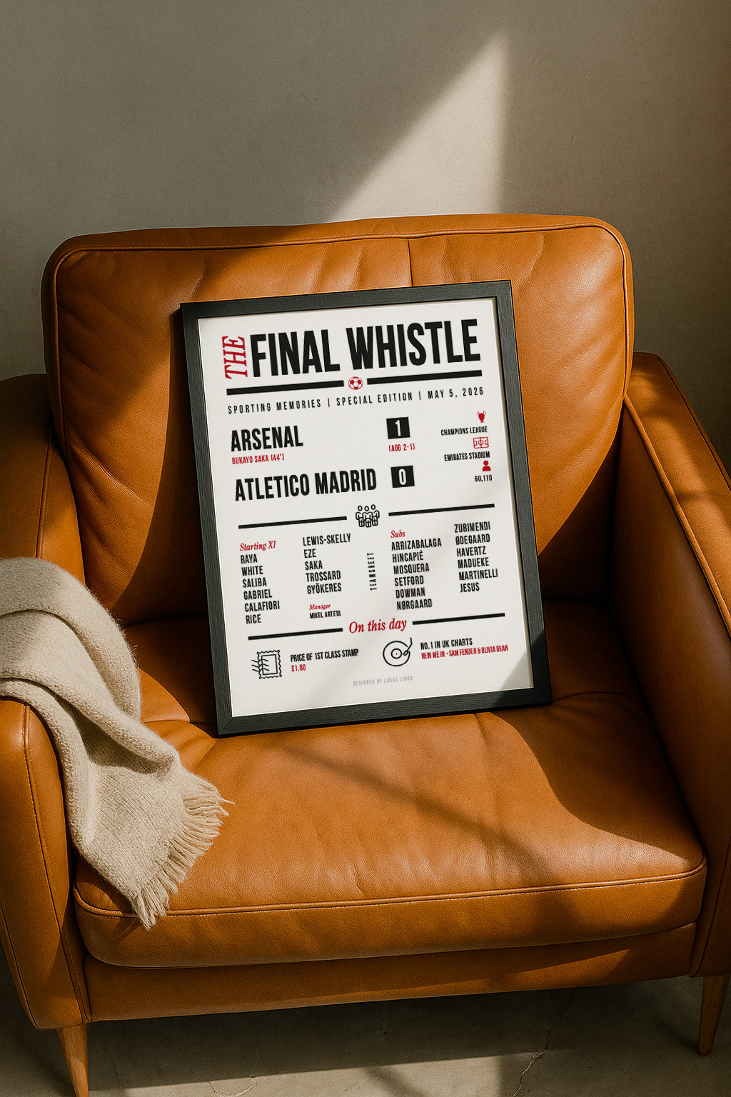 Framed "The Final Whistle" Arsenal vs Atletico Madrid Champions League match report wall art resting casually on a tan leather armchair next to a folded beige throw blanket, illuminated by warm natural sunlight. Red and black typography print for football fans, designed by Local Lingo.