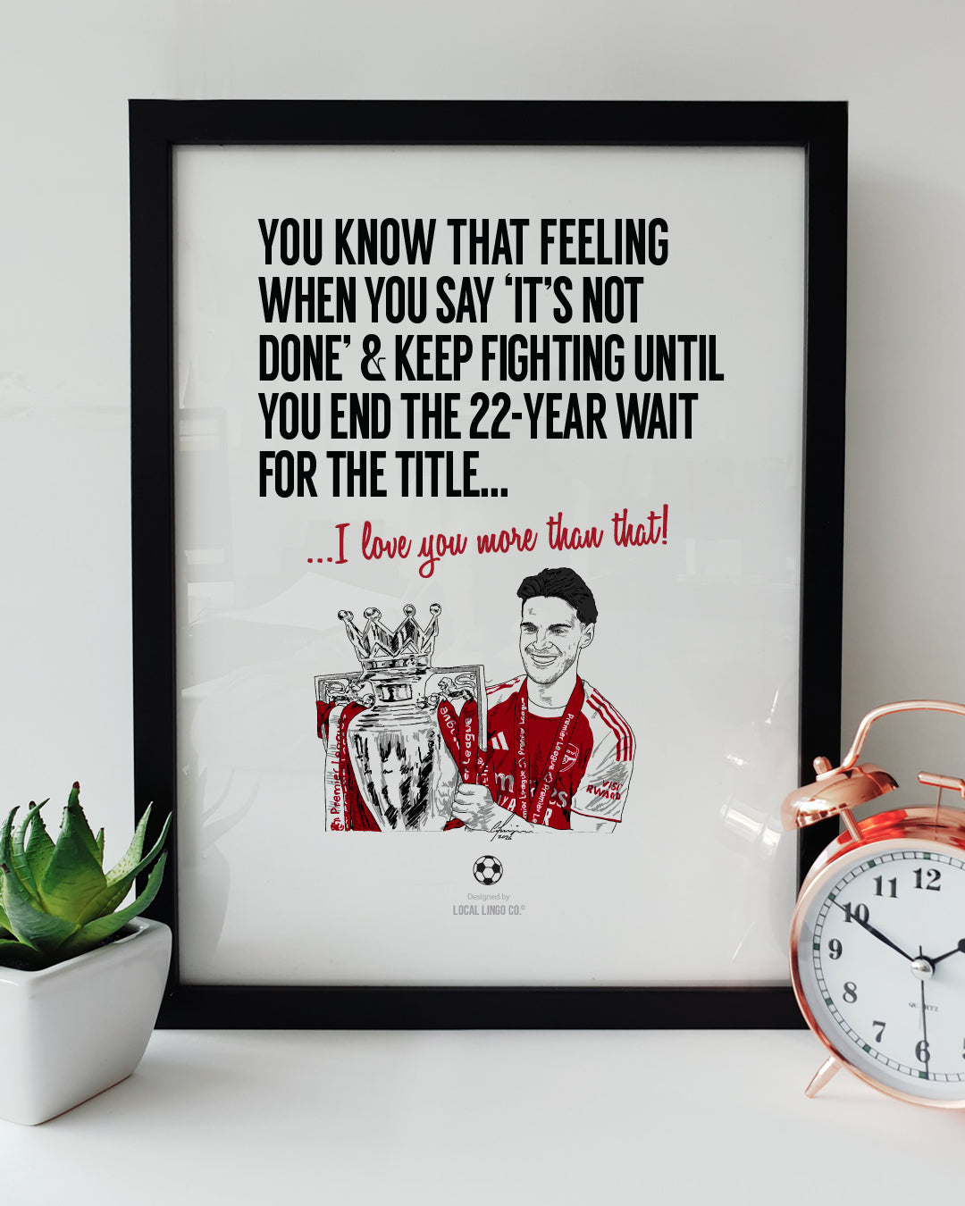 A framed football art print resting on a white surface next to a small green potted plant and a classic copper alarm clock. Bold black text reads "YOU KNOW THAT FEELING WHEN YOU SAY 'IT'S NOT DONE' & KEEP FIGHTING UNTIL YOU END THE 22-YEAR WAIT FOR THE TITLE..." followed by red text reading "...I love you more than that!". Below is a hand drawn illustration of Arsenal player Declan Rice holding the Premier League trophy in his red and white kit. Arsenal football memorabilia designed by Local Lingo.
