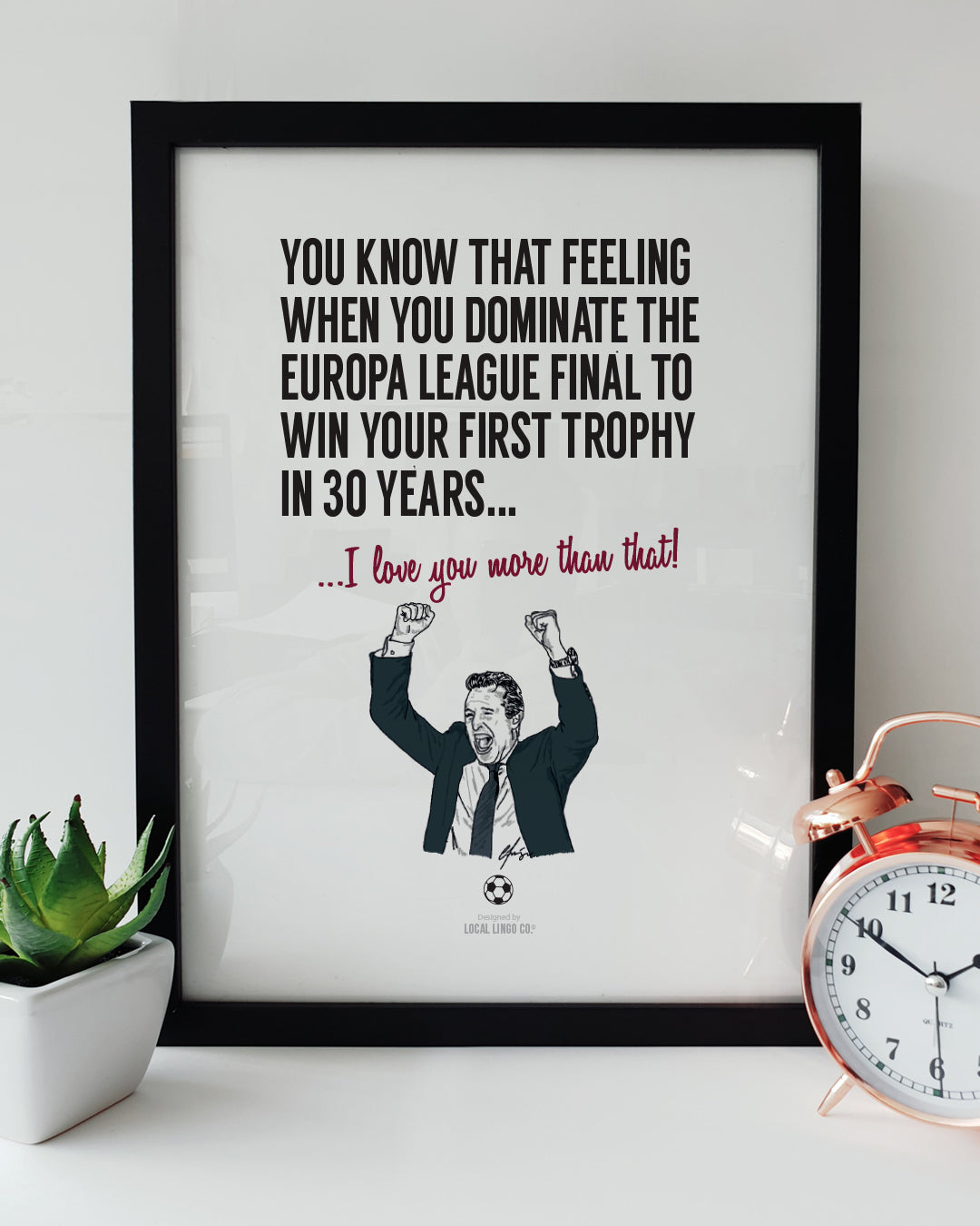 A framed football art print resting on a white surface next to a small green potted plant and a classic copper alarm clock. Bold black text reads "YOU KNOW THAT FEELING WHEN YOU DOMINATE THE EUROPA LEAGUE FINAL TO WIN YOUR FIRST TROPHY IN 30 YEARS..." followed by claret text reading "...I love you more than that!". Below is a hand-drawn illustration of Aston Villa manager Unai Emery celebrating with his fists raised. Aston Villa football memorabilia designed by Local Lingo.