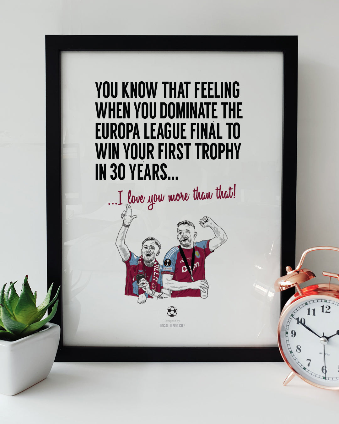 A framed football art print resting on a white surface next to a small green potted plant and a classic copper alarm clock. Bold black text reads "YOU KNOW THAT FEELING WHEN YOU DOMINATE THE EUROPA LEAGUE FINAL TO WIN YOUR FIRST TROPHY IN 30 YEARS..." followed by claret red text reading "...I love you more than that!". Below is a hand drawn illustration of Aston Villa players Matty Cash and John McGinn passionately celebrating their victory in claret and blue kits. Aston Villa football memorabilia designed 