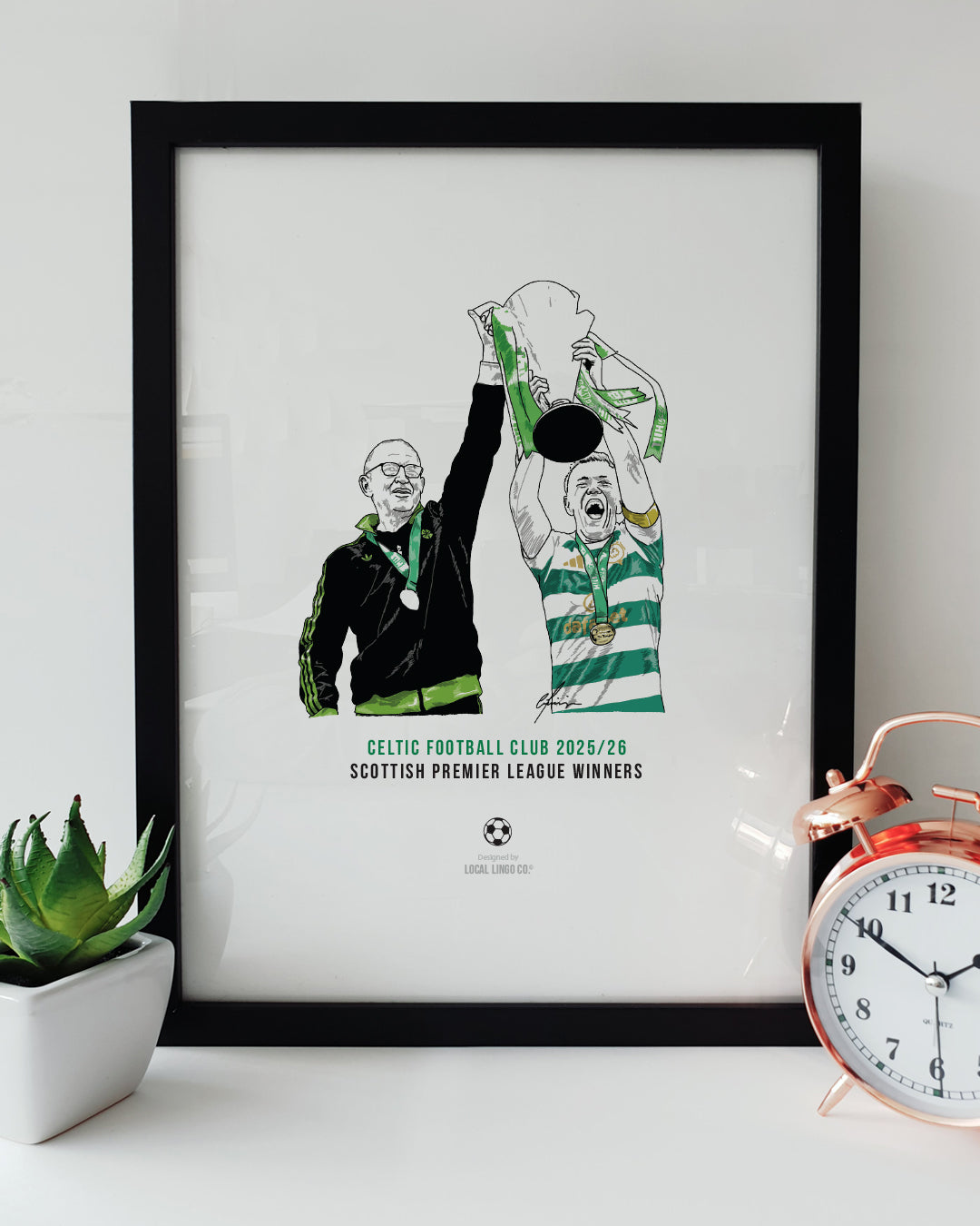 A framed football art print celebrating Celtic's 2025/26 Scottish Premiership title win. The design features a hand-drawn illustration of Martin O'Neill and Callum McGregor lifting the league trophy, displayed on a white surface next to a small green potted plant and a classic copper alarm clock. Modern Celtic football memorabilia designed by Local Lingo.
