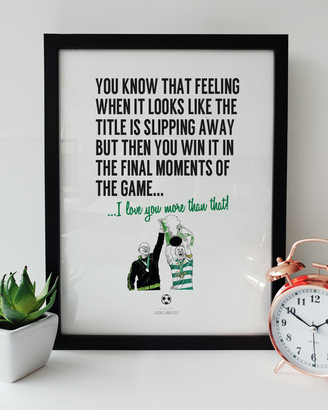 A framed football art print resting on a white surface next to a small green potted plant and a classic copper alarm clock. Bold black text reads "YOU KNOW THAT FEELING WHEN IT LOOKS LIKE THE TITLE IS SLIPPING AWAY BUT THEN YOU WIN IT IN THE FINAL MOMENTS OF THE GAME..." followed by green text reading "...I love you more than that!". Below is a hand-drawn illustration of Martin O'Neill and Callum McGregor lifting the league trophy. Celtic football memorabilia designed by Local Lingo.