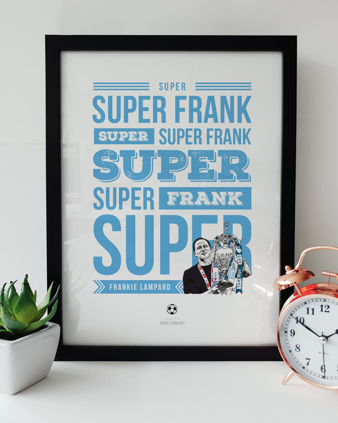 Framed typography print featuring the Coventry City fan chant "SUPER SUPER FRANK" in bold, varying sky blue text. Includes a black and white illustration of manager Frank Lampard lifting the Championship trophy. Displayed on a clean white shelf next to a copper alarm clock and a small potted succulent. Designed by Local Lingo.