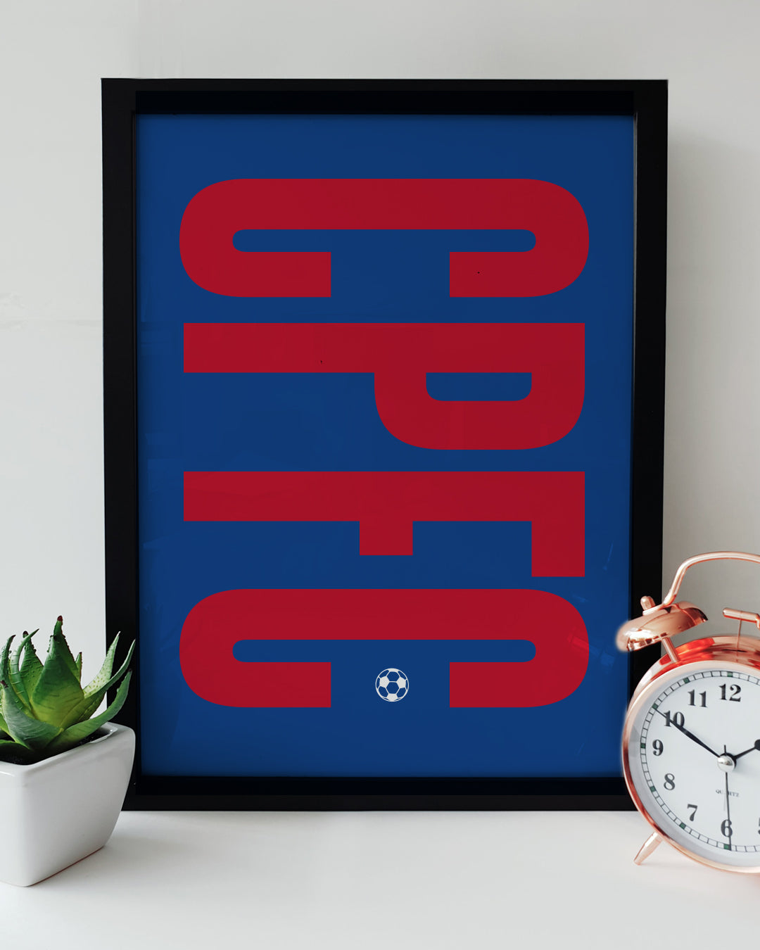 A framed CPFC typography art print resting on a white surface next to a small green potted plant and a classic copper alarm clock. The print features bold, retro-style red 'CPFC' lettering stretching vertically across a solid blue background, with a small white football icon at the base. Modern Crystal Palace football decor designed by Local Lingo.