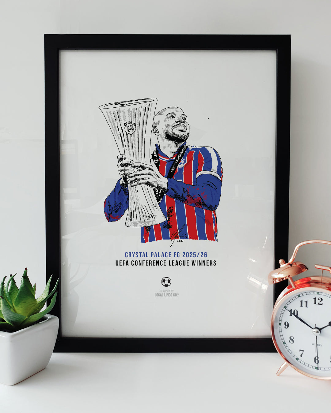 A framed football art print resting on a white surface next to a small green potted plant and a classic copper alarm clock. The print features a hand drawn illustration of Crystal Palace striker Jean-Philippe Mateta lifting the UEFA Conference League trophy. Mateta's shirt is coloured in vibrant red and blue stripes. Below the illustration, bold text reads "CRYSTAL PALACE FC 2025/26" in blue and "UEFA CONFERENCE LEAGUE WINNERS" in black. Crystal Palace football memorabilia designed by Local Lingo.