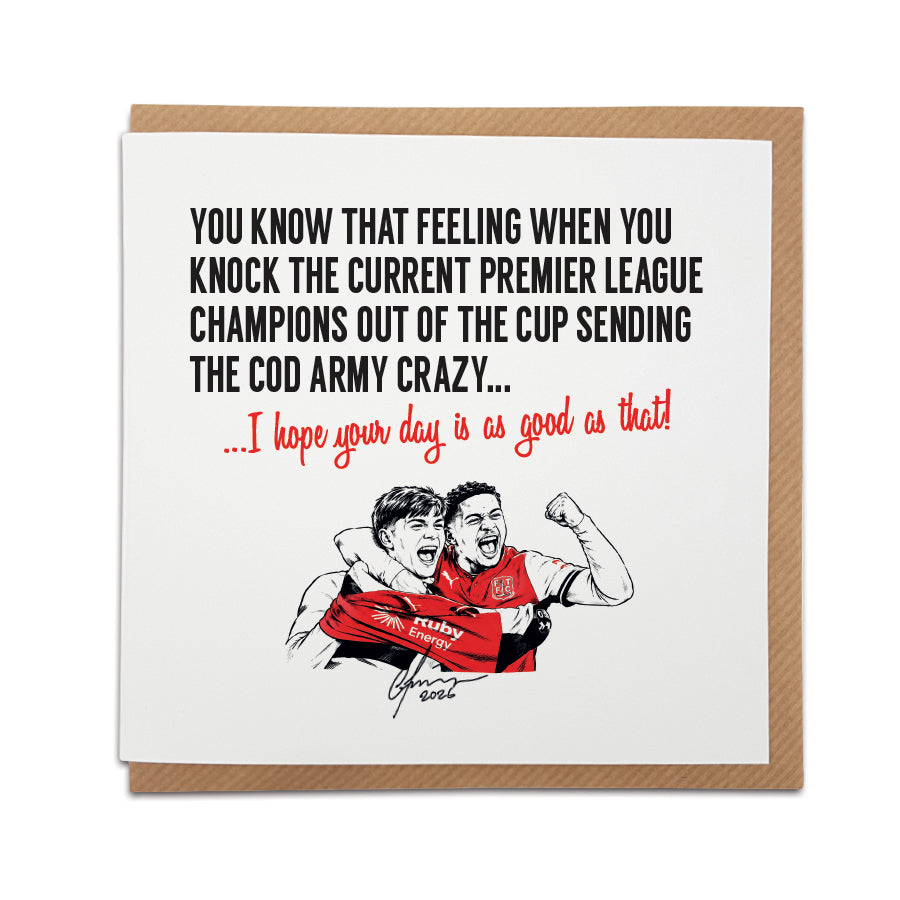 Square greeting card resting on a brown kraft envelope. Bold black text reads "YOU KNOW THAT FEELING WHEN YOU KNOCK THE CURRENT PREMIER LEAGUE CHAMPIONS OUT OF THE CUP SENDING THE COD ARMY CRAZY..." followed by red cursive text reading "...I hope your day is as good as that!". Below is a hand-drawn illustration of two Fleetwood Town players celebrating in red shirts.