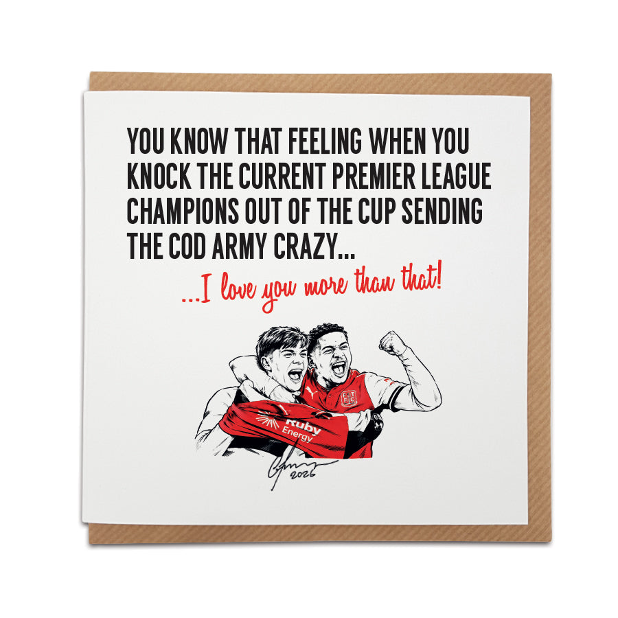 Square greeting card resting on a brown kraft envelope. Bold black text reads "YOU KNOW THAT FEELING WHEN YOU KNOCK THE CURRENT PREMIER LEAGUE CHAMPIONS OUT OF THE CUP SENDING THE COD ARMY CRAZY..." followed by red cursive text reading "...I love you more than that!". Below is a hand-drawn illustration of two Fleetwood Town players celebrating in red shirts