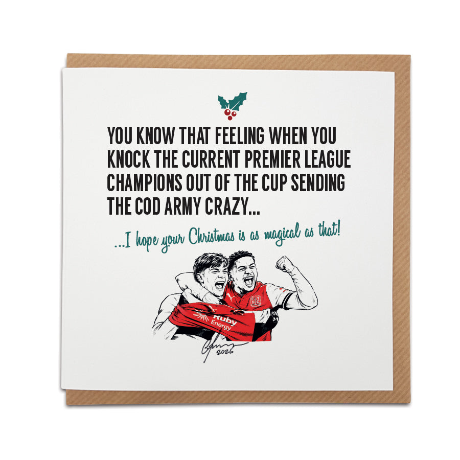 Square Christmas card resting on a brown kraft envelope. A small holly leaf graphic sits above bold black text reading "YOU KNOW THAT FEELING WHEN YOU KNOCK THE CURRENT PREMIER LEAGUE CHAMPIONS OUT OF THE CUP SENDING THE COD ARMY CRAZY..." followed by festive green cursive text reading "...I hope your Christmas is as magical as that!". Below is a hand-drawn illustration of two Fleetwood Town players celebrating in red shirts.