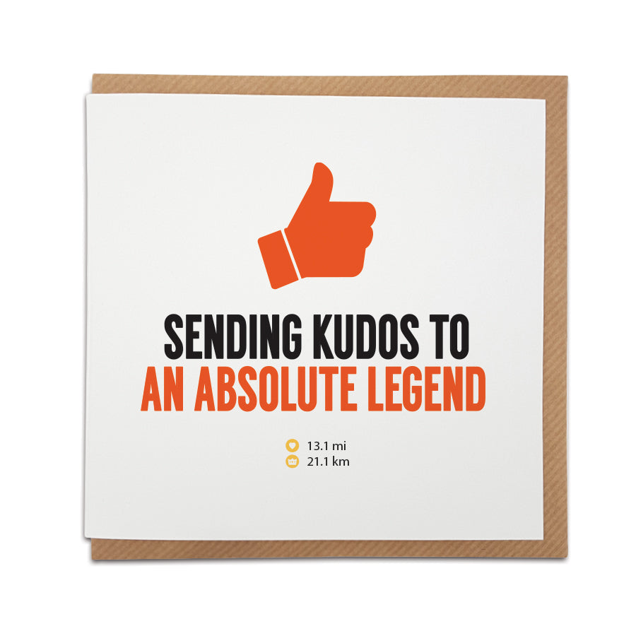 Square congratulations card resting on a brown kraft envelope. The design features a large orange thumbs-up icon above bold text reading "SENDING KUDOS TO" in black and "AN ABSOLUTE LEGEND" in orange. Small text below displays the half marathon distances "13.1 mi" and "21.1 km". Funny running themed greeting card designed by Local Lingo.