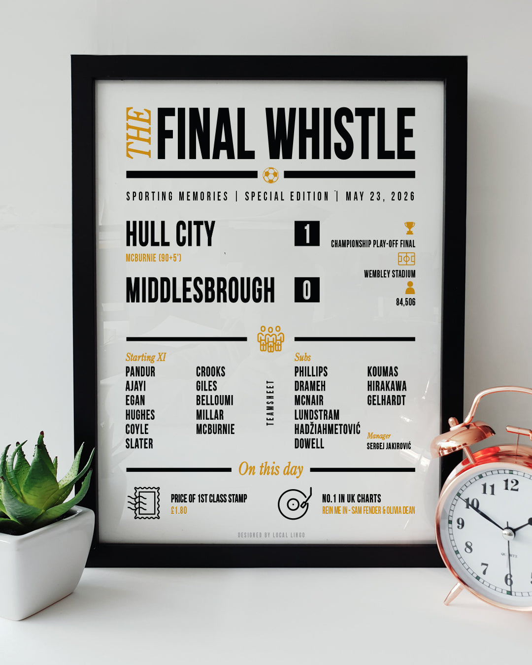 A framed 'Final Whistle' football keepsake print celebrating Hull City's 2026 Championship Play-Off Final 1-0 victory over Middlesbrough at Wembley Stadium. The print features the match stats, starting lineup, and "On this day" trivia in amber and black text. It is displayed on a white surface next to a small green potted plant and a classic copper alarm clock. Modern football memorabilia designed by Local Lingo.