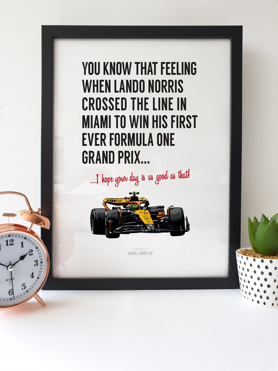 Framed artwork featuring a quote about Lando Norris winning his first Formula 1 Grand Prix in Miami, with an illustration of his McLaren F1 car. The text reads, "You know that feeling when Lando Norris crossed the line in Miami to win his first ever Formula One Grand Prix... I hope your day is as good as that!" The artwork is placed on a white desk next to a copper alarm clock and a small potted plant. designed by local lingo