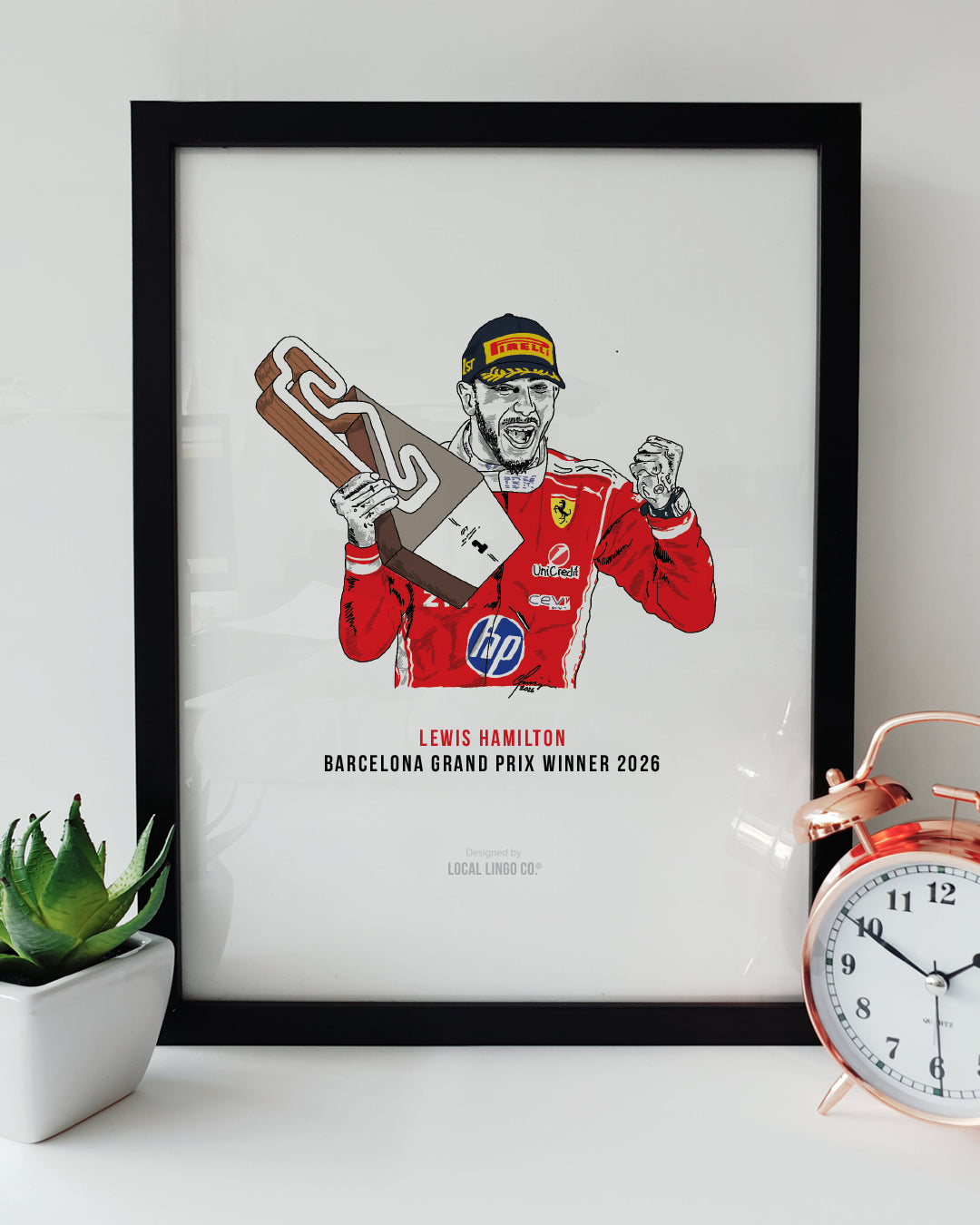 A framed Formula 1 art print resting on a white surface next to a small green potted plant and a classic copper alarm clock. The print features a hand-drawn illustration of F1 driver Lewis Hamilton celebrating with a unique trophy in his red Ferrari race suit. Below the illustration, bold text reads "LEWIS HAMILTON" in red and "BARCELONA GRAND PRIX WINNER 2026" in black. Lewis Hamilton Formula 1 print designed by Local Lingo.