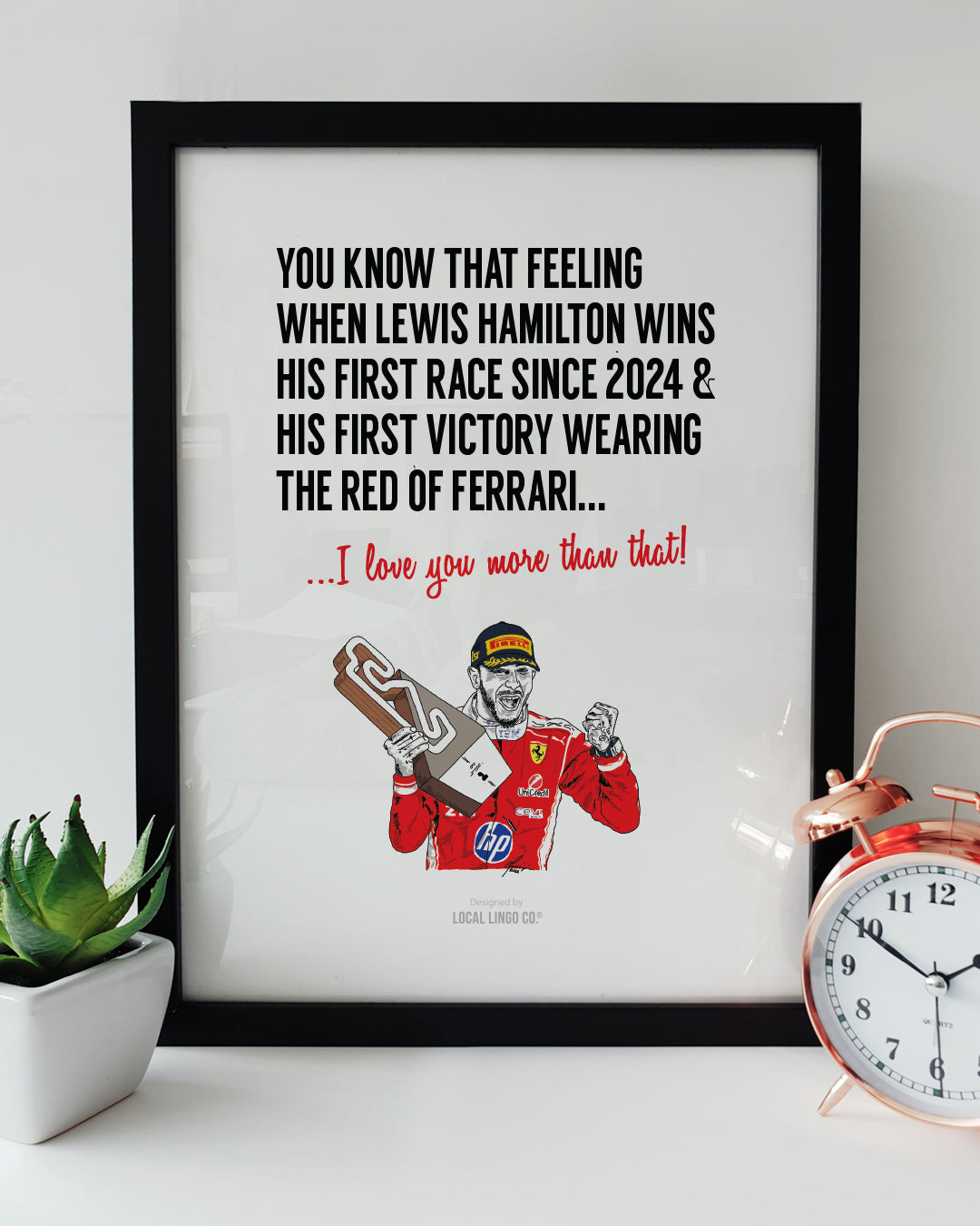 A framed Formula 1 art print resting on a white surface next to a small green potted plant and a classic copper alarm clock. Bold black text reads "YOU KNOW THAT FEELING WHEN LEWIS HAMILTON WINS HIS FIRST RACE SINCE 2024 & HIS FIRST VICTORY WEARING THE RED OF FERRARI..." followed by red text reading "...I love you more than that!". Below is a hand-drawn illustration of F1 driver Lewis Hamilton celebrating with a trophy in his red Ferrari race suit. Lewis Hamilton Formula 1 print designed by Local Lingo.