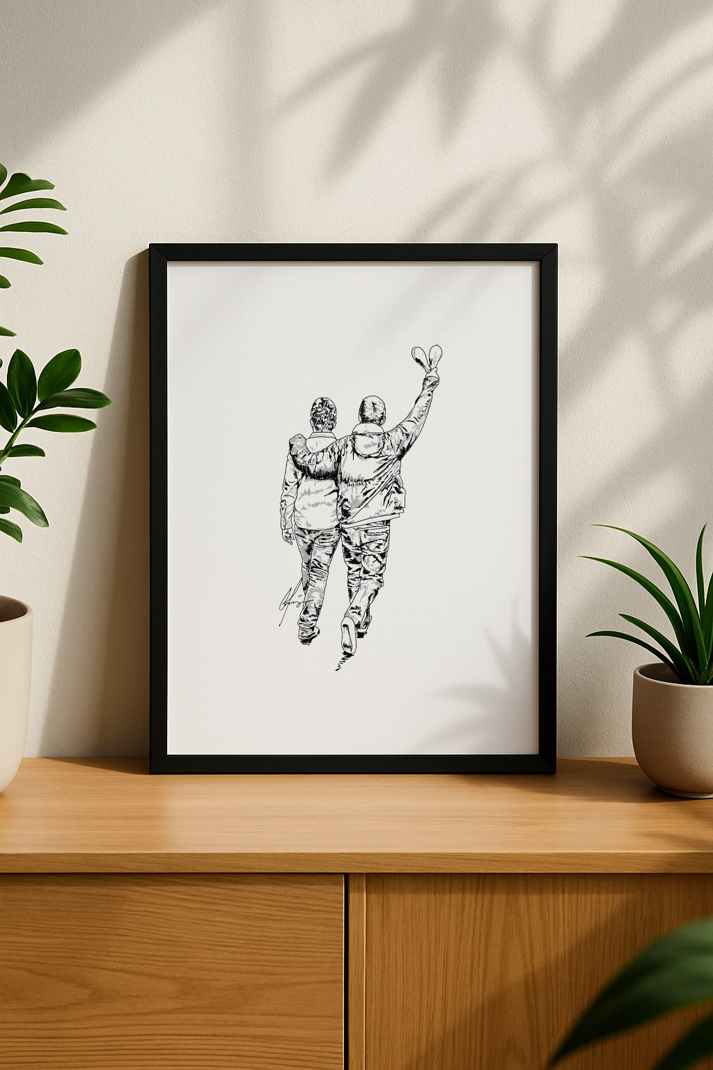 Framed black and white art print on a wooden cabinet. The artwork features a hand-drawn sketch of Liam and Noel Gallagher walking away with their arms around each other. Minimalist Oasis reunion fan art by Local Lingo.