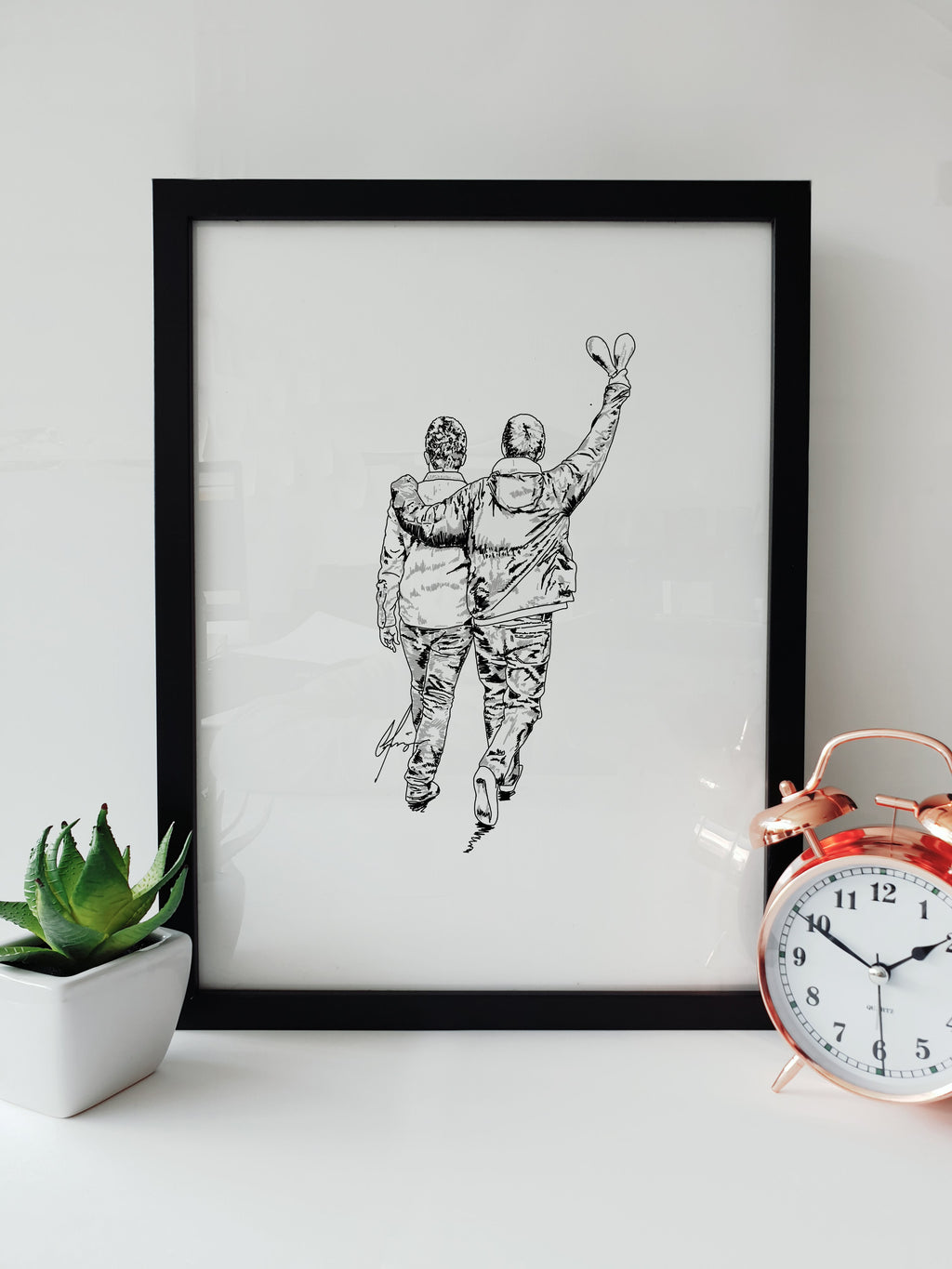 Framed black and white art print on a white shelf. The artwork features a hand-drawn sketch of Liam and Noel Gallagher walking away with their arms around each other. Minimalist Oasis reunion fan art by Local Lingo.