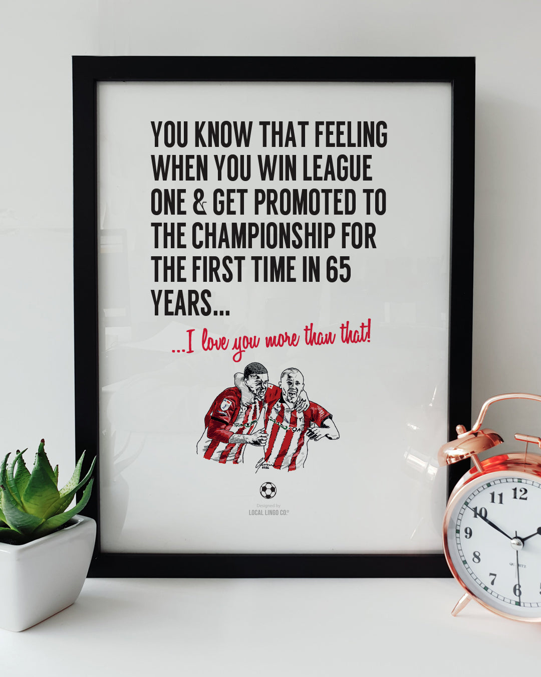 Framed football wall art celebrating Lincoln City's promotion. Bold black text reads: "You know that feeling when you win League One & get promoted to the Championship for the first time in 65 years...". Below it, red script reads "...I love you more than that!". Features a hand-drawn illustration of two Lincoln City football players embracing and celebrating in red and white striped kits. Displayed in a black frame next to a green succulent in a white pot and a copper alarm clock. Sentimental Lincoln City 