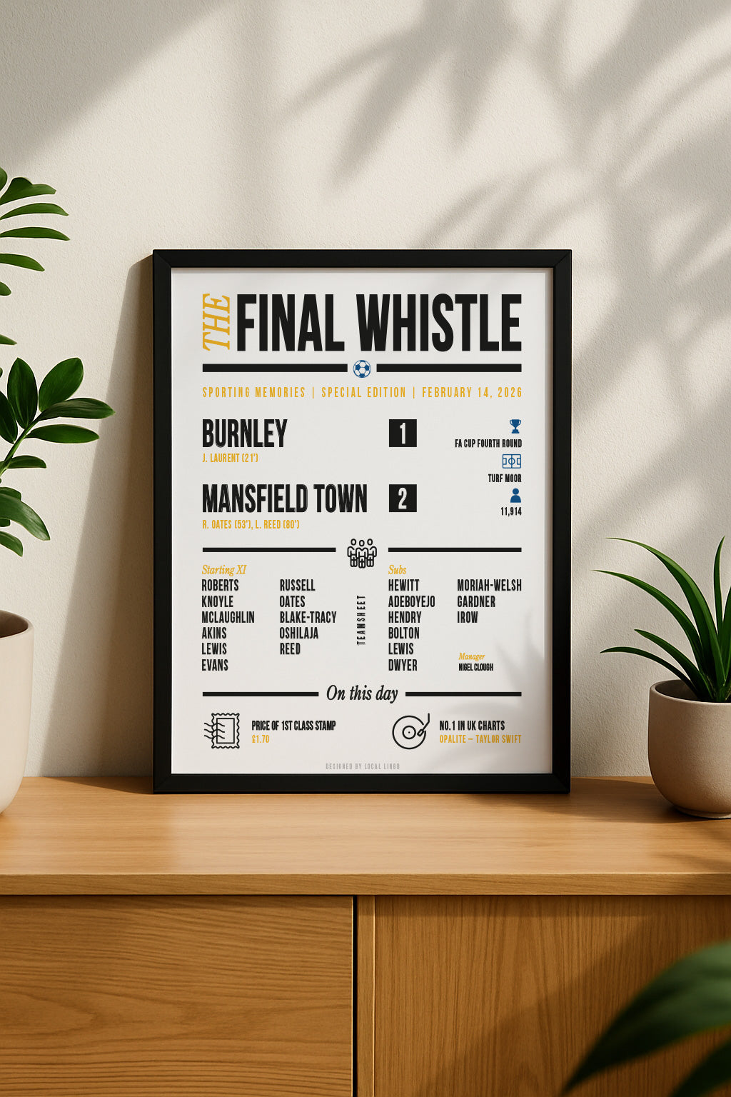 Vertical framed poster of the Mansfield Town vs Burnley match report print hanging on a beige wall. The bold black text and yellow/blue accents stand out against the clean white background, styled above a wooden sideboard with green plants for a modern home interior look. Designed by Local Lingo.