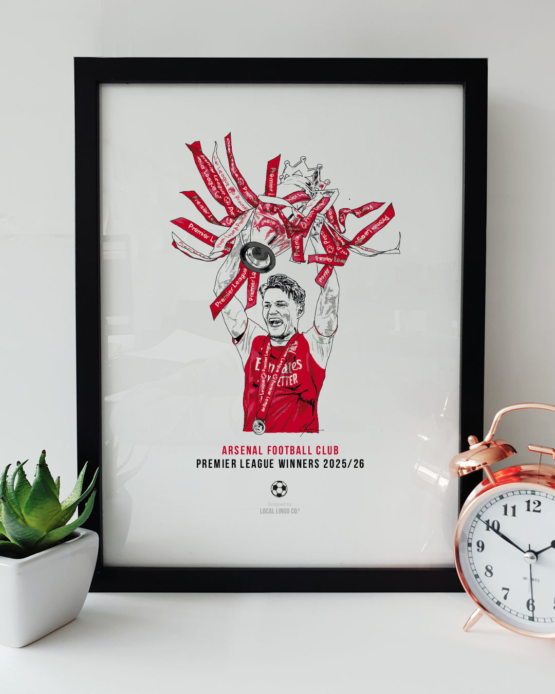 A framed football art print resting on a white surface next to a small green potted plant and a classic copper alarm clock. The print features a hand drawn illustration of Arsenal captain Martin Odegaard lifting the Premier League trophy above his head. The trophy ribbons and Odegaard's shirt are coloured in a vibrant red. Below the illustration, bold text reads "ARSENAL FOOTBALL CLUB PREMIER LEAGUE WINNERS 2025/26" in red and black. Arsenal football memorabilia designed by Local Lingo.