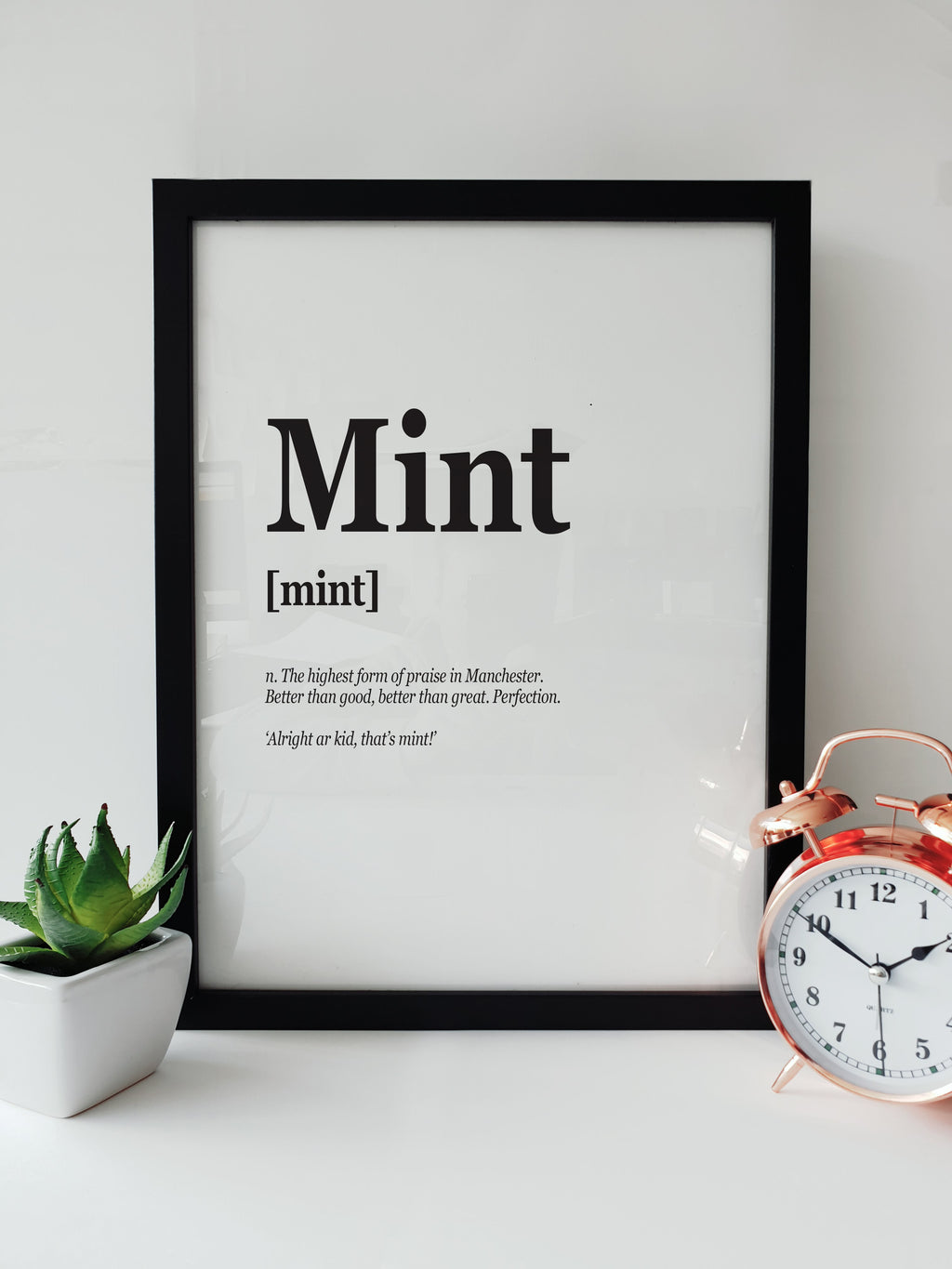 Framed typographic print on a white shelf titled "Mint". The text defines the term as "The highest form of praise in Manchester. Better than good, better than great. Perfection." Displayed next to a small plant and a copper alarm clock. Designed by Local Lingo.