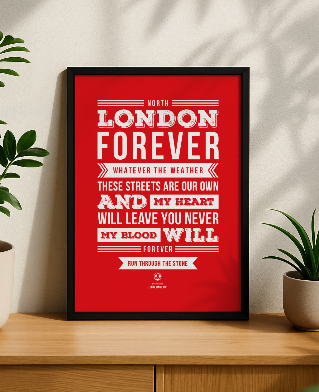 A framed football art print displayed vertically on a light wooden sideboard, flanked by two small green potted plants with natural sunlight casting shadows on the wall behind it. The print features the Arsenal 'North London Forever' chant lyrics by Louis Mark Dunford in bold vintage style white typography on a solid red background. Arsenal football wall art designed by Local Lingo.