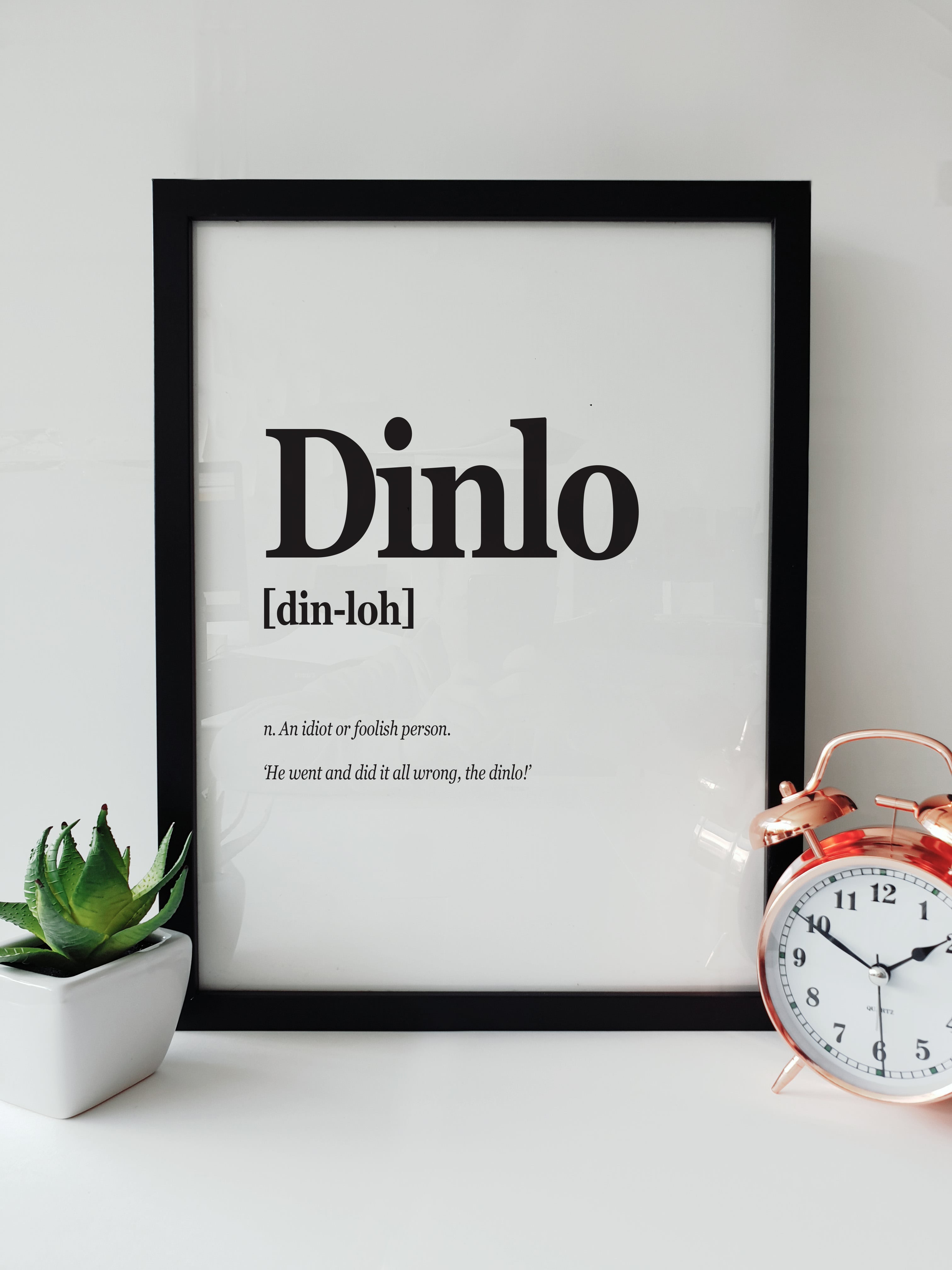 Framed Portsmouth Dialect Print featuring the word "Dinlo" with pronunciation and definition, displayed on a white desk with a plant and a copper alarm clock. noun translation poster designed by local lingo
