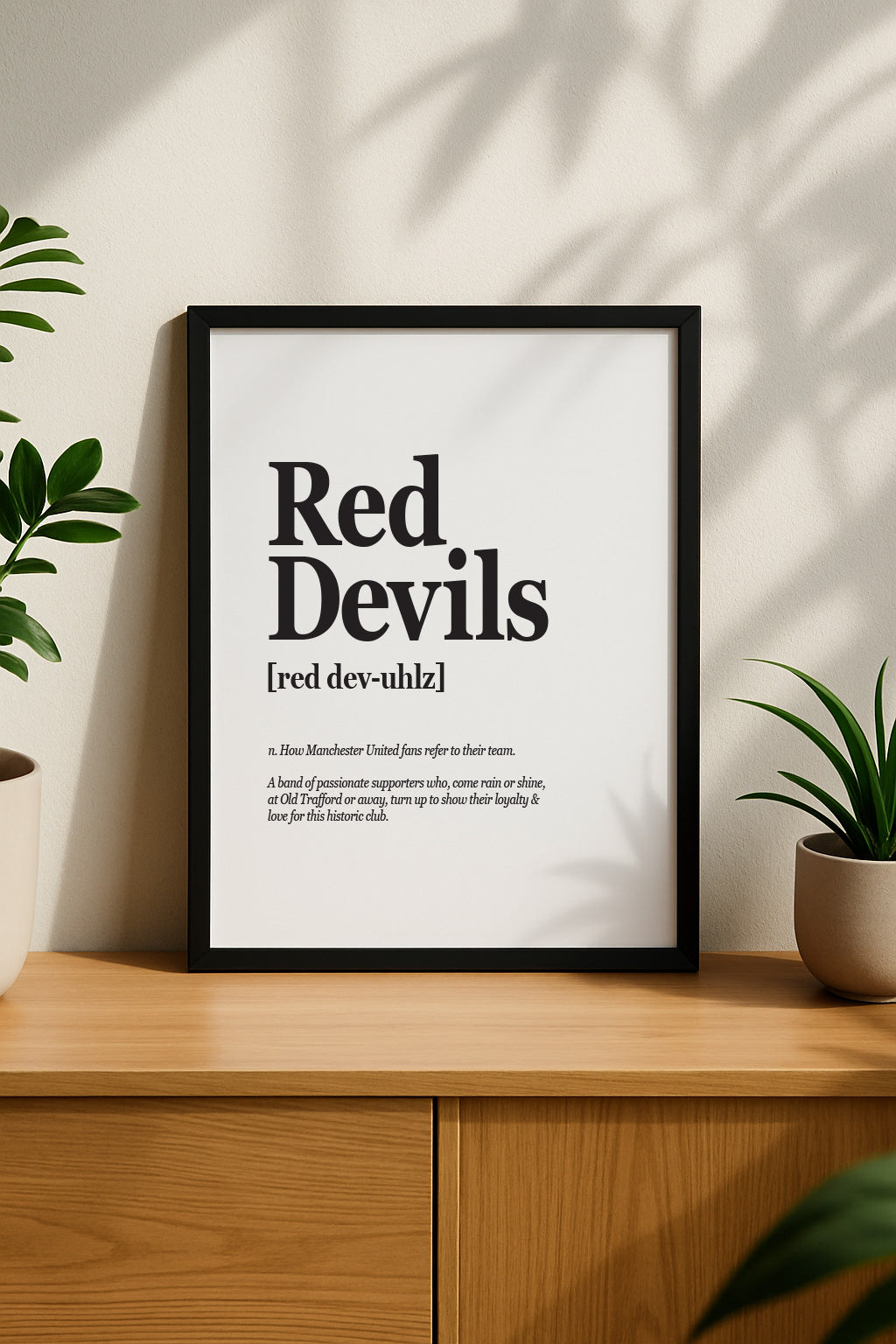 Vertical framed poster of the "Red Devils" definition print hanging on a beige wall. The bold black text stands out against the clean white background, styled above a wooden sideboard with green plants for a modern home interior look. Designed by Local Lingo.