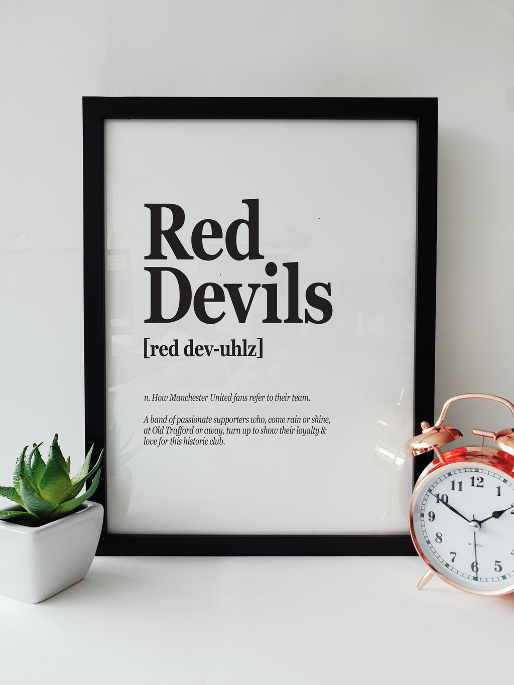 Framed typographic print on a white shelf titled "Red Devils". The text defines the term as "How Manchester United fans refer to their team" and "A band of passionate supporters." Displayed next to a small plant and a copper alarm clock. Designed by Local Lingo.