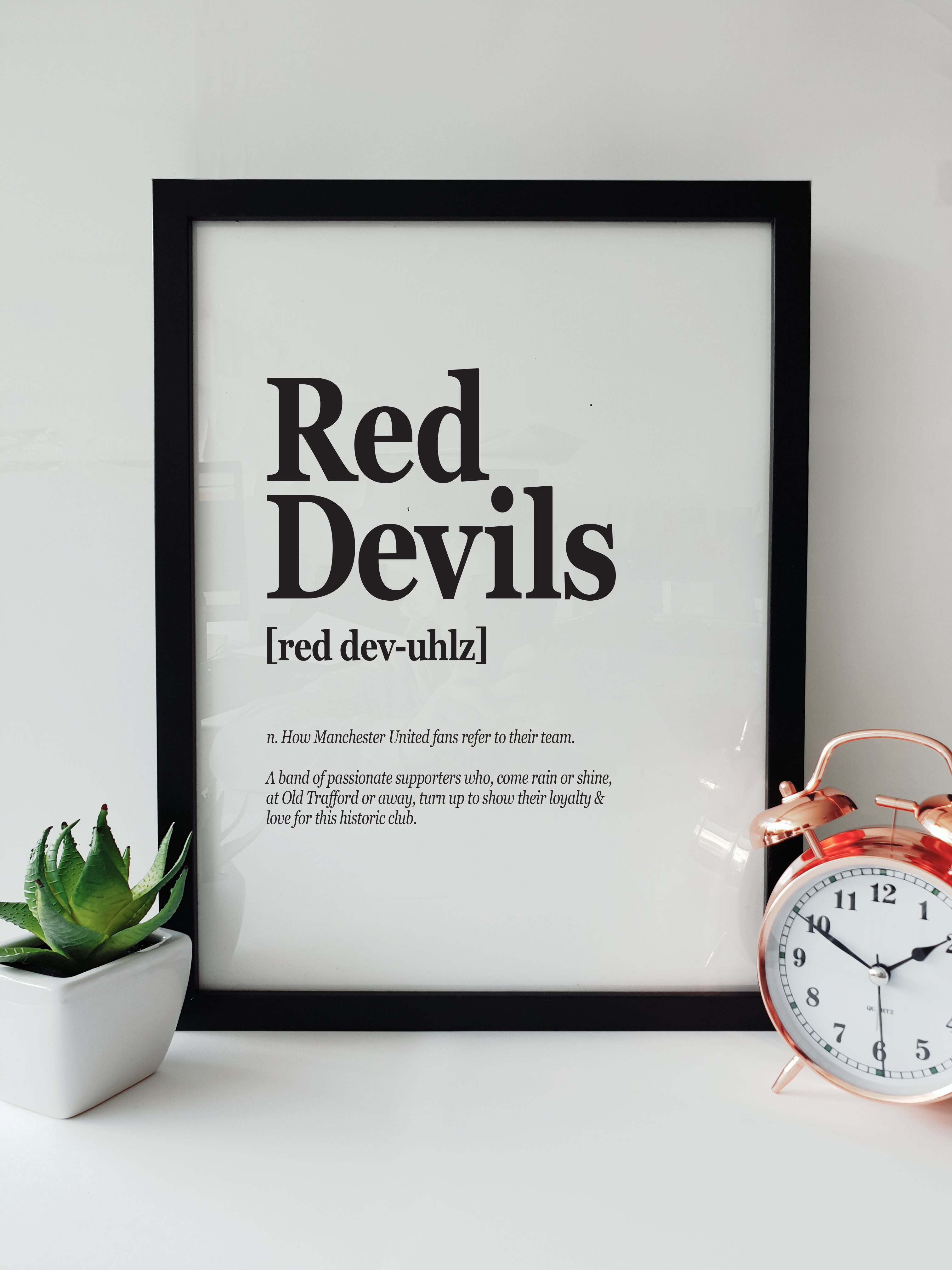 Framed typographic print on a white shelf titled "Red Devils". The text defines the term as "How Manchester United fans refer to their team" and "A band of passionate supporters." Displayed next to a small plant and a copper alarm clock. Designed by Local Lingo.