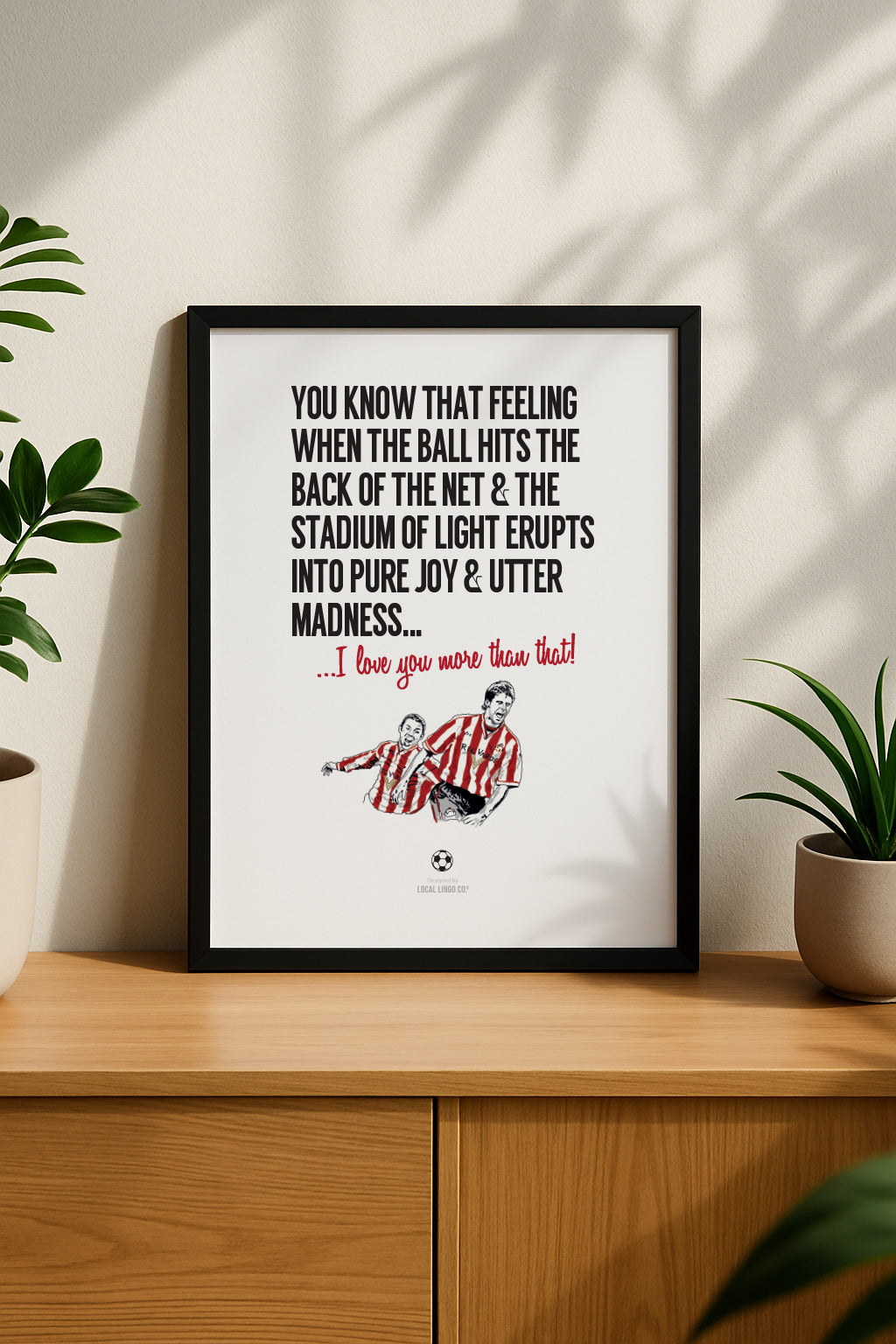 Vertical framed art print for Sunderland AFC fans resting on a modern wooden sideboard next to green house plants. The poster features a funny romantic football quote about the Stadium of Light and a red and white player illustration. Designed by Local Lingo.