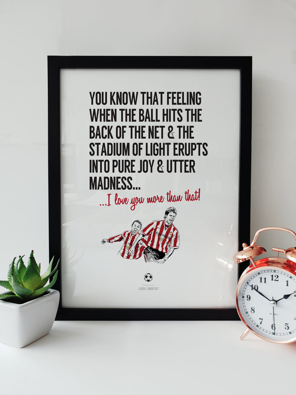Framed typographic print on a white surface next to a copper alarm clock and a small potted plant. The text reads: "You know that feeling when the ball hits the back of the net & the Stadium of Light erupts into pure joy & utter madness... I love you more than that!". Features a hand-drawn illustration of Sunderland players celebrating. Designed by Local Lingo.