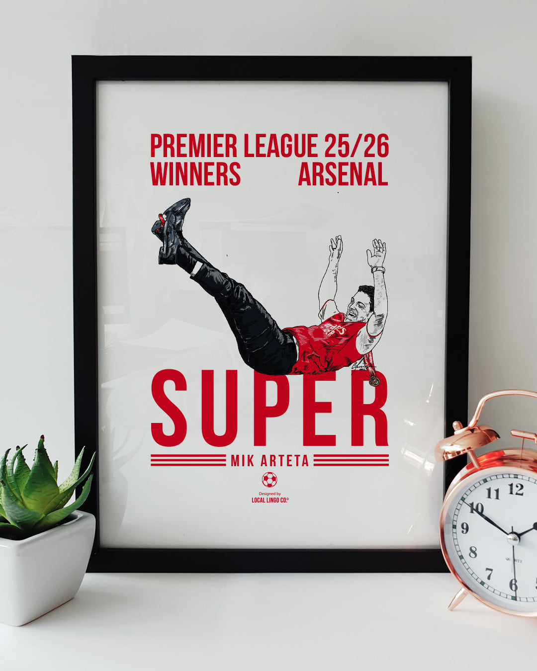 A framed football art print resting on a white surface next to a small green potted plant and a classic copper alarm clock. The print features a magazine cover style design with bold red text reading "PREMIER LEAGUE 25/26 WINNERS ARSENAL" at the top and "SUPER MIK ARTETA" at the bottom. In the centre is a hand drawn illustration of Arsenal manager Mikel Arteta wildly celebrating, leaning back with his legs kicked up in the air, wearing a red shirt and black trousers. Arsenal football memorabilia designed by