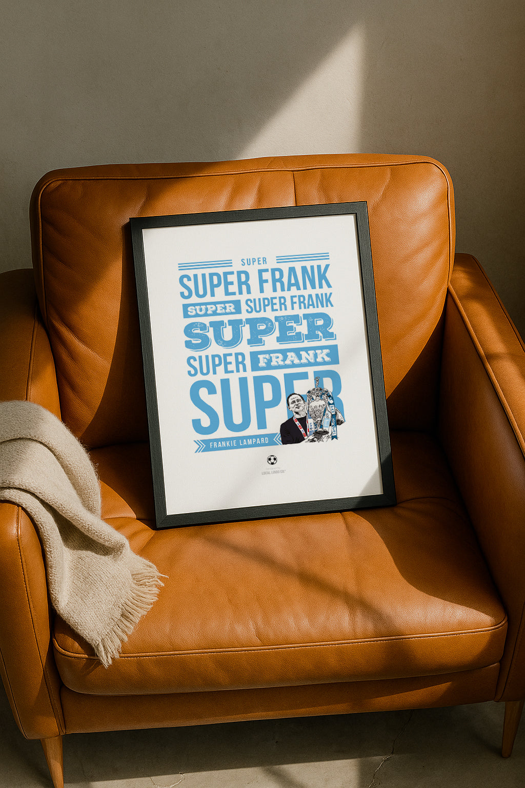 Framed "Super Frankie Lampard" Coventry City fan chant wall art resting casually on a tan leather armchair next to a folded beige throw blanket, illuminated by warm natural sunlight. Sky blue typography print for football fans, designed by Local Lingo.