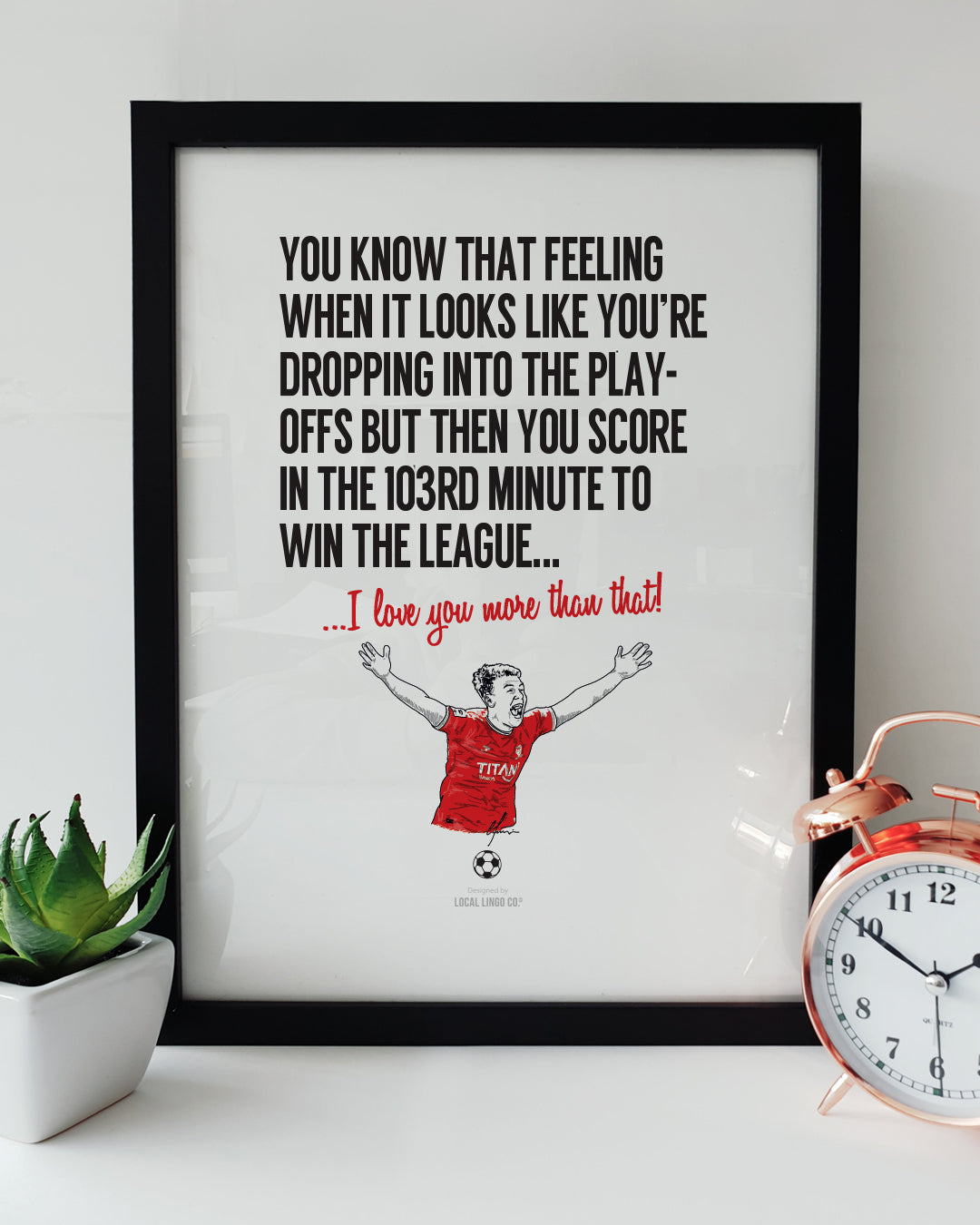 Framed football wall art celebrating York City's 103rd-minute league-winning goal. Bold black text reads: "You know that feeling when it looks like you're dropping into the play-offs but then you score in the 103rd minute to win the league...". Below it, red script reads "...I love you more than that!". Features a hand-drawn illustration of a York City football player celebrating with arms raised in a red kit. Displayed in a black frame next to a green succulent in a white pot and a copper alarm clock. Sent